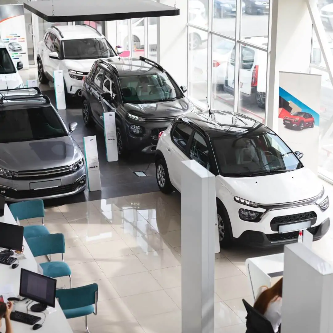 High-angle view of a car dealership showroom with polished white tile floors, featuring various clean cars and blue chairs at consultation desks.