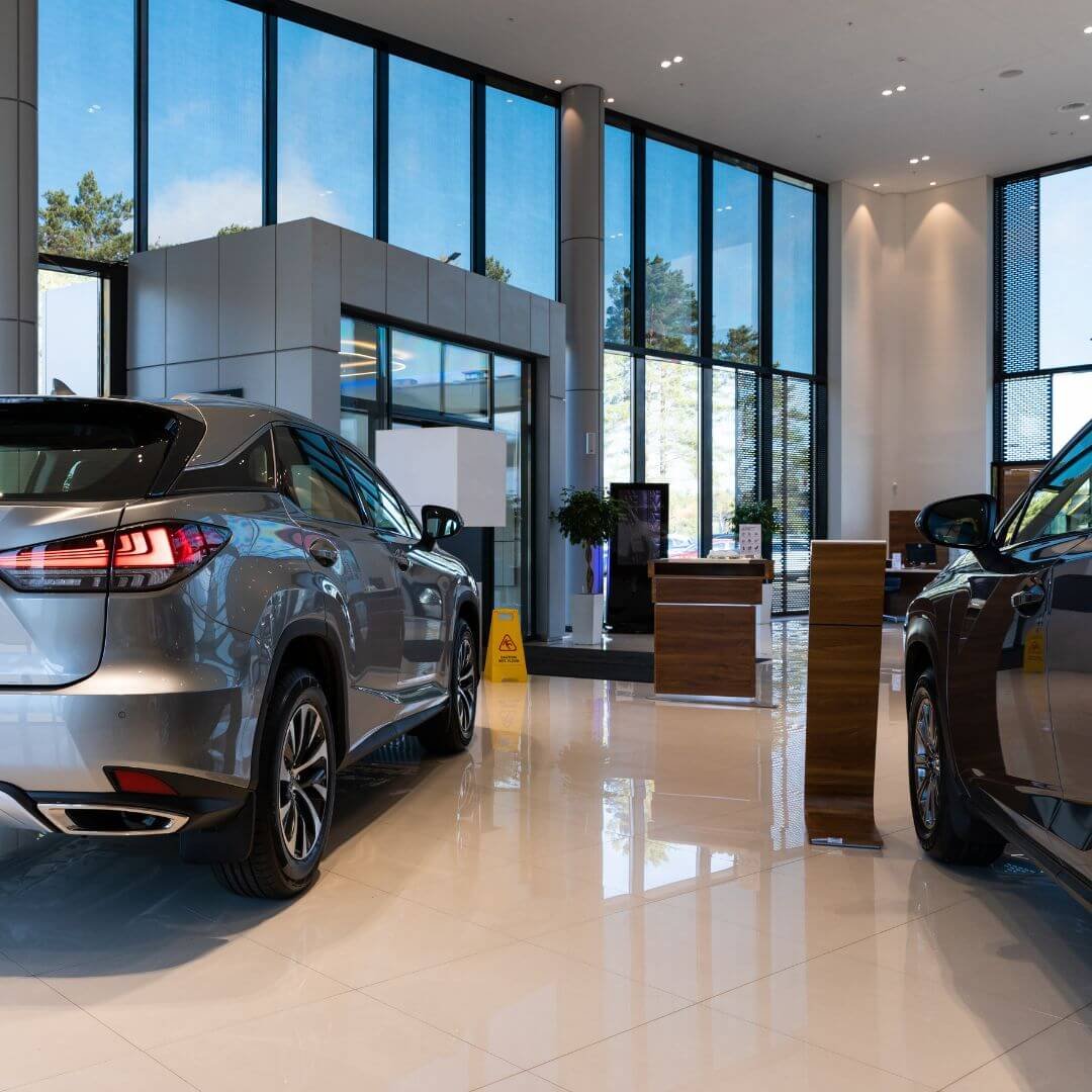 Interior of a luxury car dealership with highly reflective floors, large floor-to-ceiling windows, and a yellow caution wet floor sign.