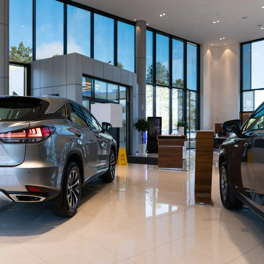 Interior of a luxury car dealership with highly reflective floors, large floor-to-ceiling windows, and a yellow caution wet floor sign.