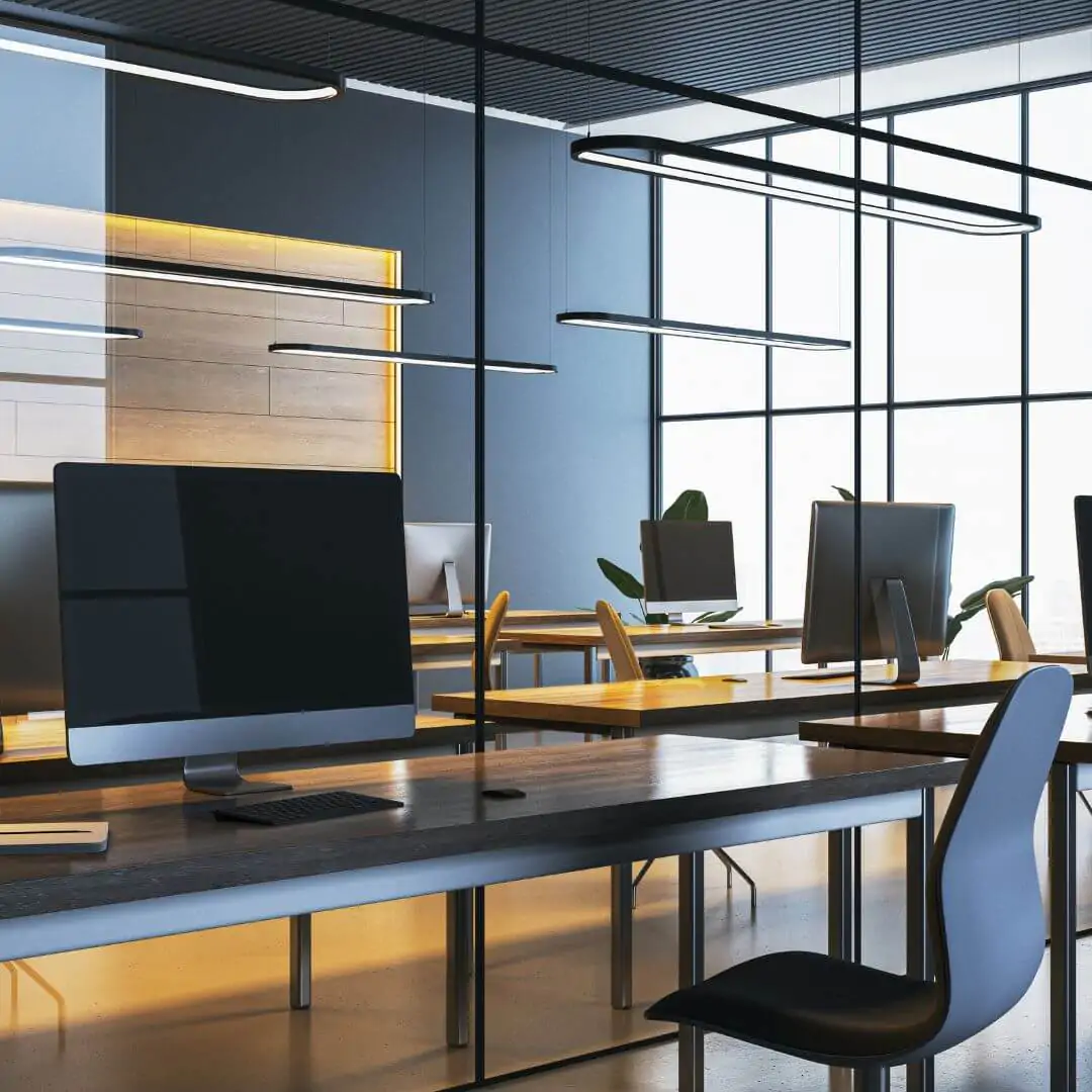 A clean modern office space featuring dark wooden desks with black monitors, glass partitions, and sleek black linear pendant lights.