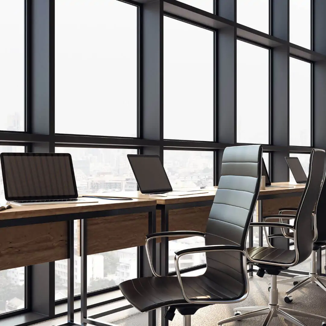Close-up of a clean office workstation featuring wooden desks, black ergonomic chairs, and laptops in front of large windows with a city view.