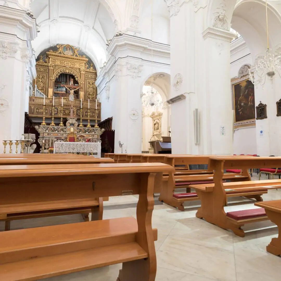 The bright interior of a grand white church featuring a highly detailed gold altar and rows of polished wooden pews.