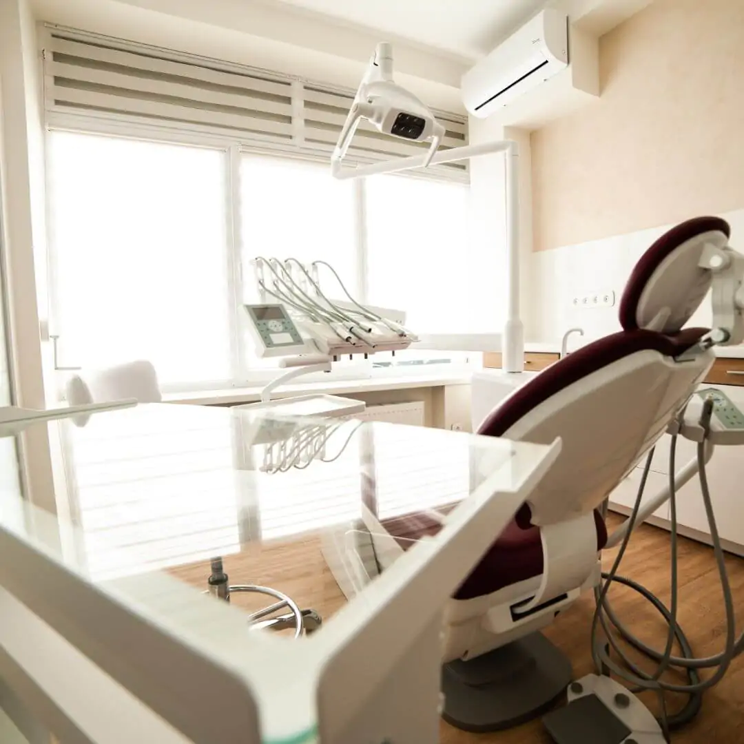 A bright and sterile dental exam room with a white chair, maroon headrest, and sanitized medical equipment near a window.