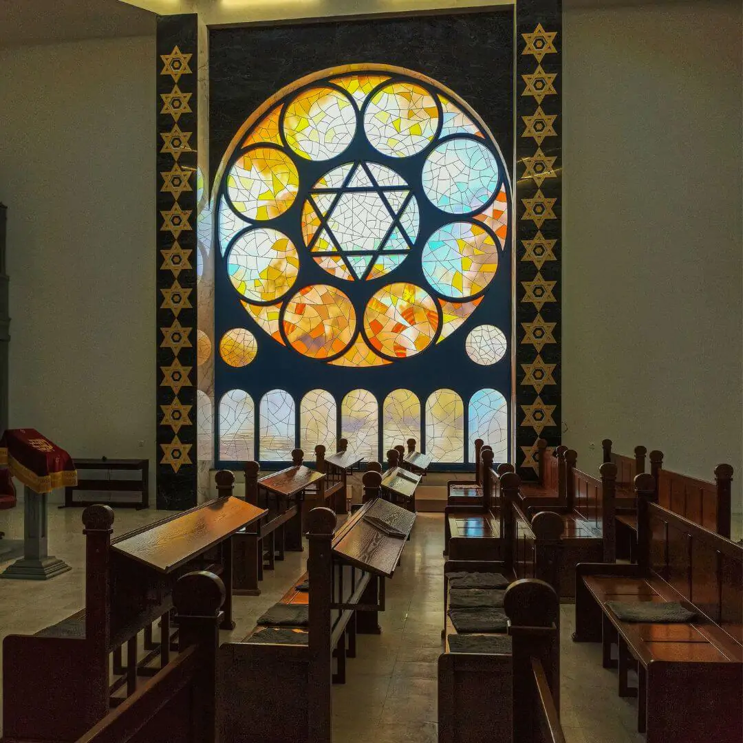 Interior of a religious space with wooden pews facing a large, colorful circular stained-glass window featuring the Star of David.