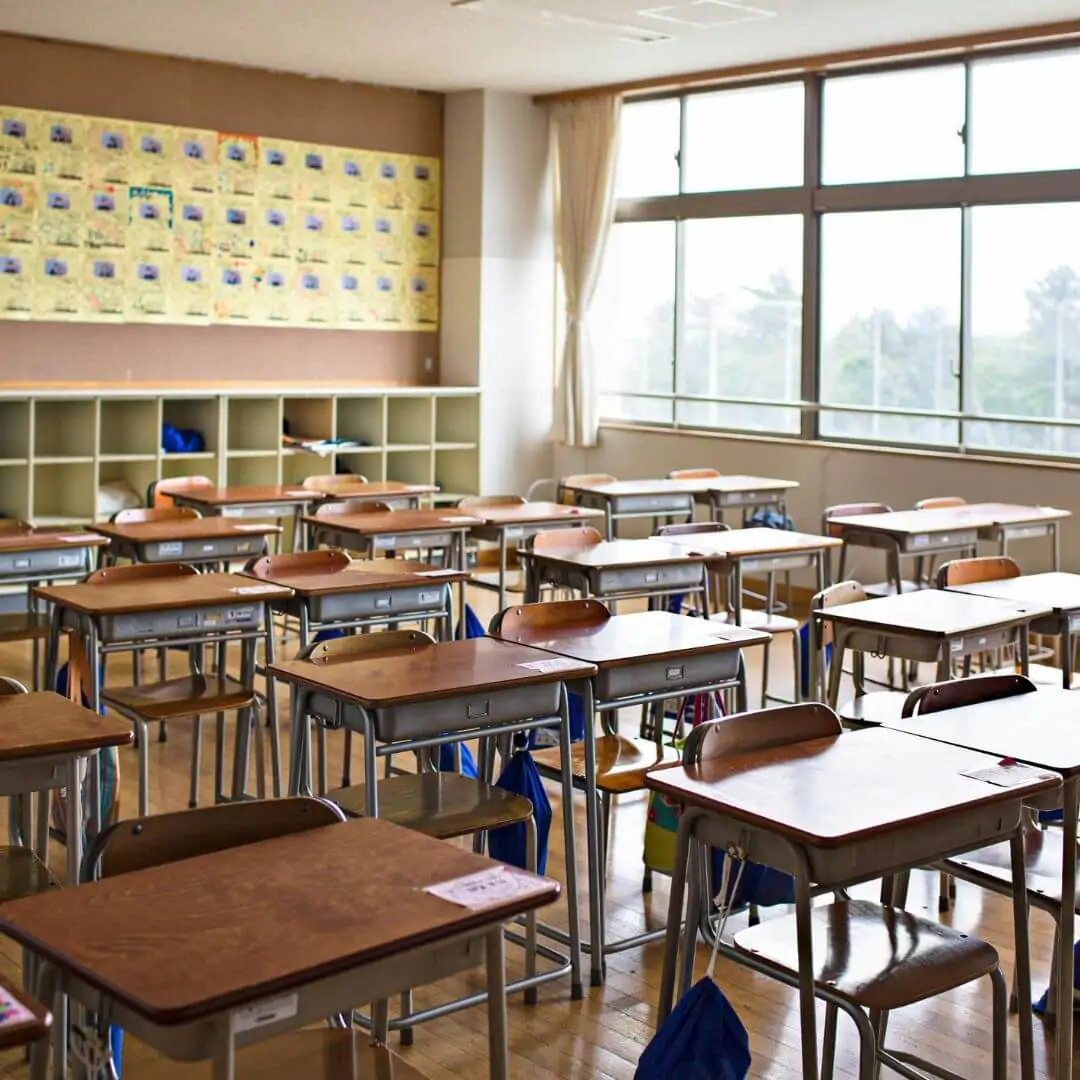 A bright, clean traditional classroom with rows of wooden desks, blue chairs, and educational posters on the wall.
